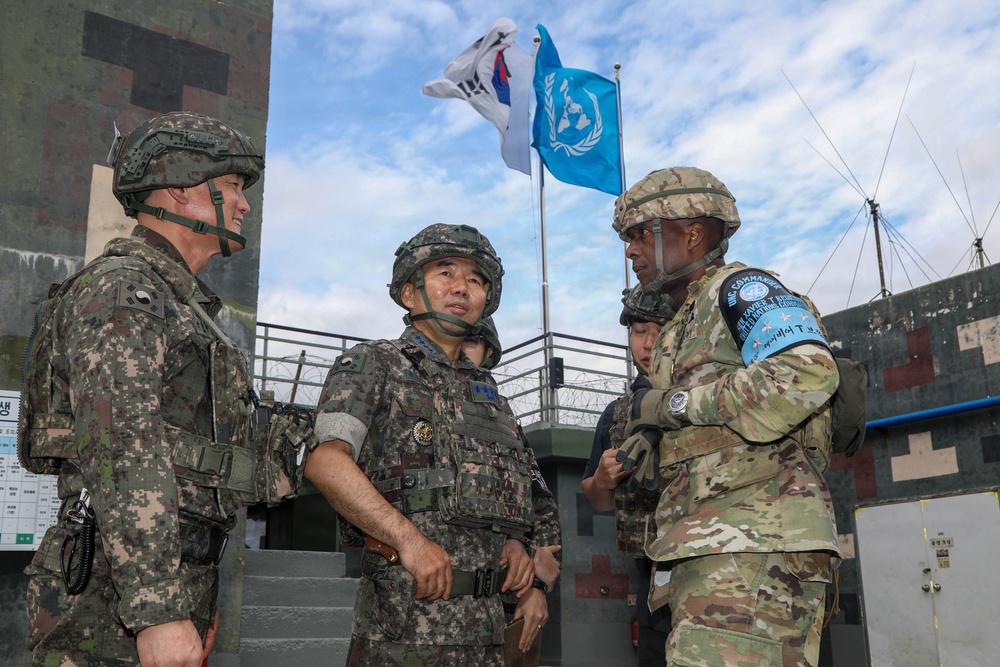 The image shows a group of military personnel in conversation at what appears to be a fortified checkpoint or observation post. Two soldiers wearing South Korean military uniforms and helmets are speaking with a soldier in US Army MultiCam uniform bearing a blue UN Command patch on the sleeve. Two flags are visible in the background: the South Korean Taegukgi and the United Nations flag.