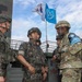 The image shows a group of military personnel in conversation at what appears to be a fortified checkpoint or observation post. Two soldiers wearing South Korean military uniforms and helmets are speaking with a soldier in US Army MultiCam uniform bearing a blue UN Command patch on the sleeve. Two flags are visible in the background: the South Korean Taegukgi and the United Nations flag.