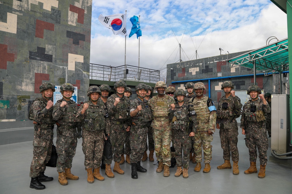 A group of military personnel pose together for a formal group photo at what appears to be a joint military installation. The group includes both South Korean Army soldiers in distinctive camouflage uniforms and what appear to be U.S. Army soldiers in MultiCam/OCP uniforms. Several individuals are making fist gestures toward the camera in a show of solidarity or camaraderie.