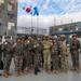 A group of military personnel pose together for a formal group photo at what appears to be a joint military installation. The group includes both South Korean Army soldiers in distinctive camouflage uniforms and what appear to be U.S. Army soldiers in MultiCam/OCP uniforms. Several individuals are making fist gestures toward the camera in a show of solidarity or camaraderie.