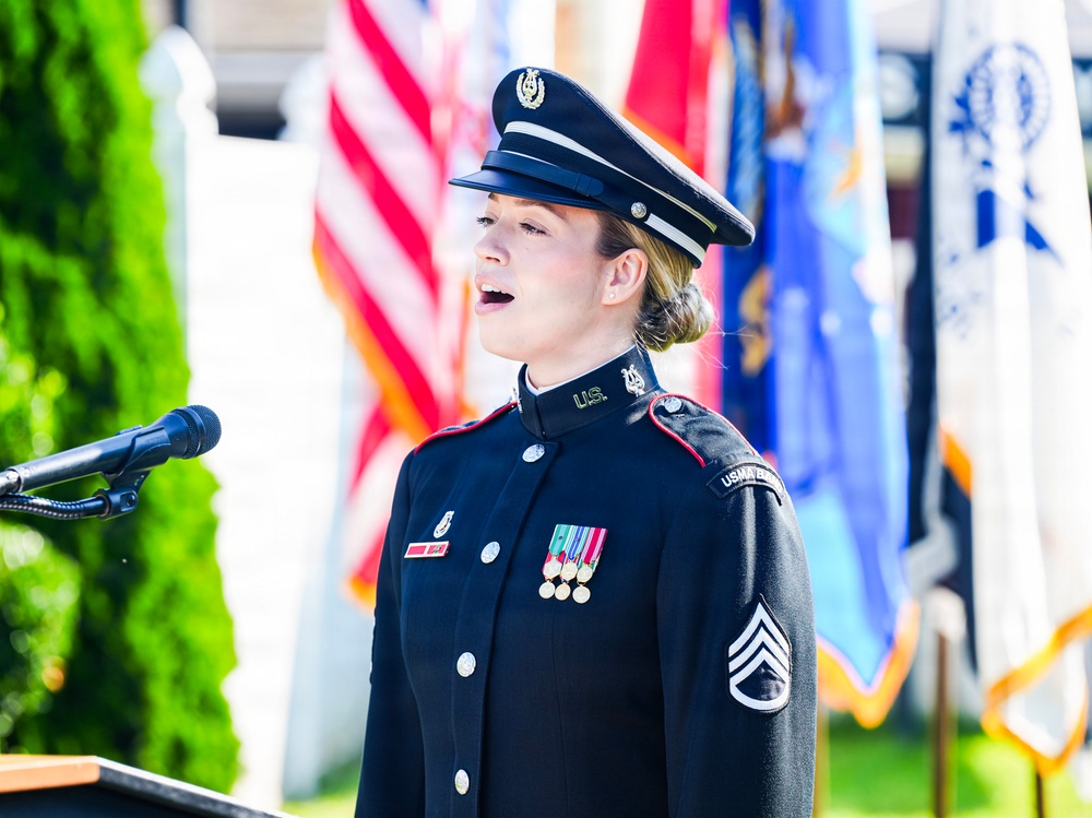 A female U.S. Army soldier in dress blues uniform is performing, likely singing, at a formal military or patriotic ceremony. She stands at a microphone podium with multiple flags visible in the background, including the American flag. Her uniform displays rank insignia of Staff Sergeant and several service ribbons/medals.