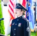 A female U.S. Army soldier in dress blues uniform is performing, likely singing, at a formal military or patriotic ceremony. She stands at a microphone podium with multiple flags visible in the background, including the American flag. Her uniform displays rank insignia of Staff Sergeant and several service ribbons/medals.