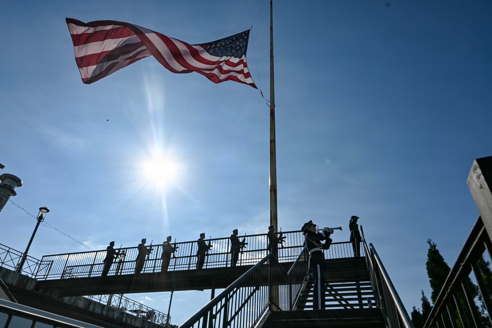 A military or ceremonial honor guard is positioned on an elevated walkway or bridge beneath a large, waving American flag on a flagpole. One figure in the foreground appears to be playing a bugle, while several others stand at attention holding rifles along the railing. The image is shot from a low angle against a bright sun and blue sky, creating a dramatic silhouette effect.