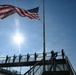 A military or ceremonial honor guard is positioned on an elevated walkway or bridge beneath a large, waving American flag on a flagpole. One figure in the foreground appears to be playing a bugle, while several others stand at attention holding rifles along the railing. The image is shot from a low angle against a bright sun and blue sky, creating a dramatic silhouette effect.