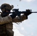 A military service member in full combat gear is actively firing an assault rifle, with a spent brass casing visibly ejecting from the weapon. The soldier is equipped with advanced tactical equipment including night vision mounts, optical sights, and a plate carrier vest. The image appears to be taken during a live-fire training exercise or operational deployment.