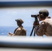 Two military personnel in desert camouflage uniforms are conducting observation operations from what appears to be a naval vessel or elevated platform overlooking open ocean. The foreground figure is using a spotting scope mounted on a tripod, while a second figure in the background wears a tactical helmet and appears to be taking notes. The setting suggests a maritime surveillance or fire support coordination exercise.