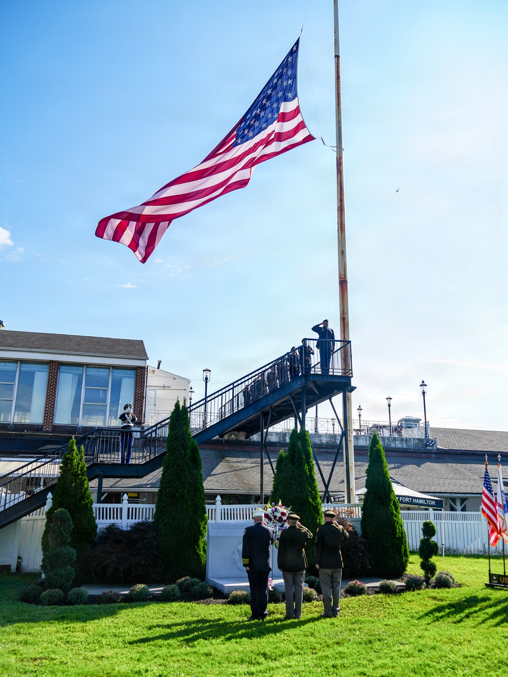 A military memorial ceremony is taking place at Fort Hamilton, visible from signage in the background. Several uniformed personnel are gathered around a memorial monument with a floral wreath, while others stand at salute on an elevated platform near a large American flag flying from a tall flagpole. The scene depicts a formal commemorative event on well-maintained grounds.