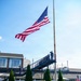 A military memorial ceremony is taking place at Fort Hamilton, visible from signage in the background. Several uniformed personnel are gathered around a memorial monument with a floral wreath, while others stand at salute on an elevated platform near a large American flag flying from a tall flagpole. The scene depicts a formal commemorative event on well-maintained grounds.