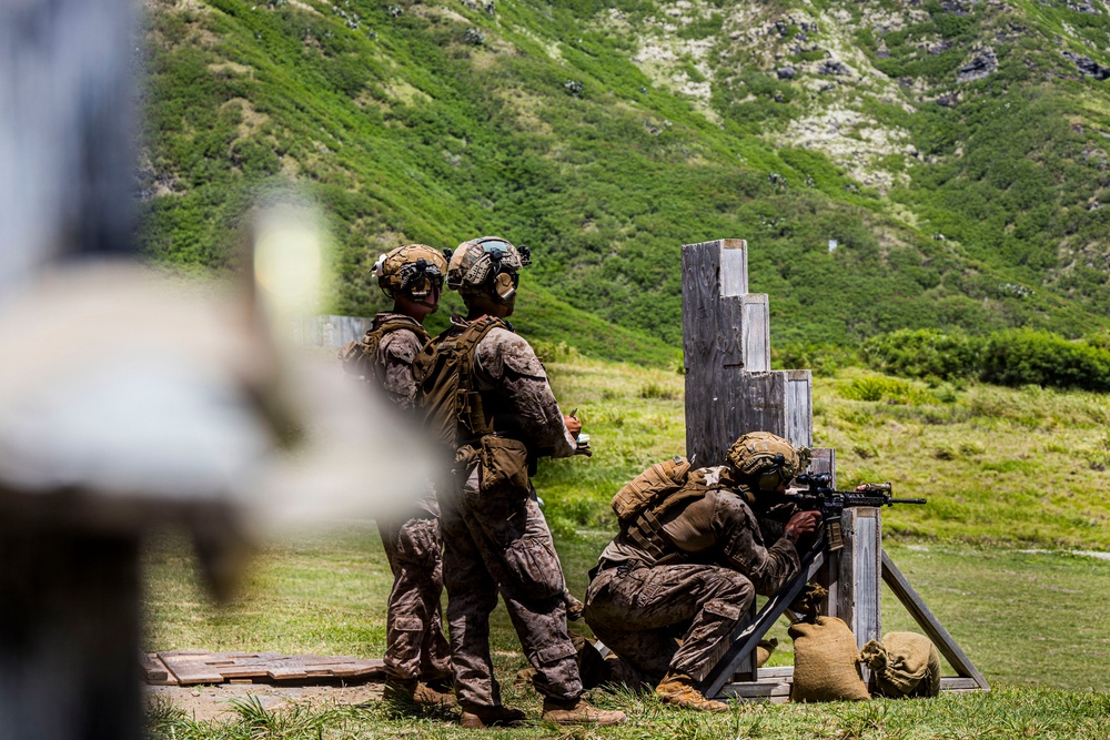 Three military personnel in camouflage uniforms and tactical gear are conducting a live-fire training exercise at an outdoor shooting range. One soldier is in a kneeling position firing a rifle from behind a wooden barricade structure, while two others stand nearby observing or instructing. The setting is a green, mountainous landscape consistent with a tropical or subtropical training environment.