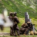 Three military personnel in camouflage uniforms and tactical gear are conducting a live-fire training exercise at an outdoor shooting range. One soldier is in a kneeling position firing a rifle from behind a wooden barricade structure, while two others stand nearby observing or instructing. The setting is a green, mountainous landscape consistent with a tropical or subtropical training environment.