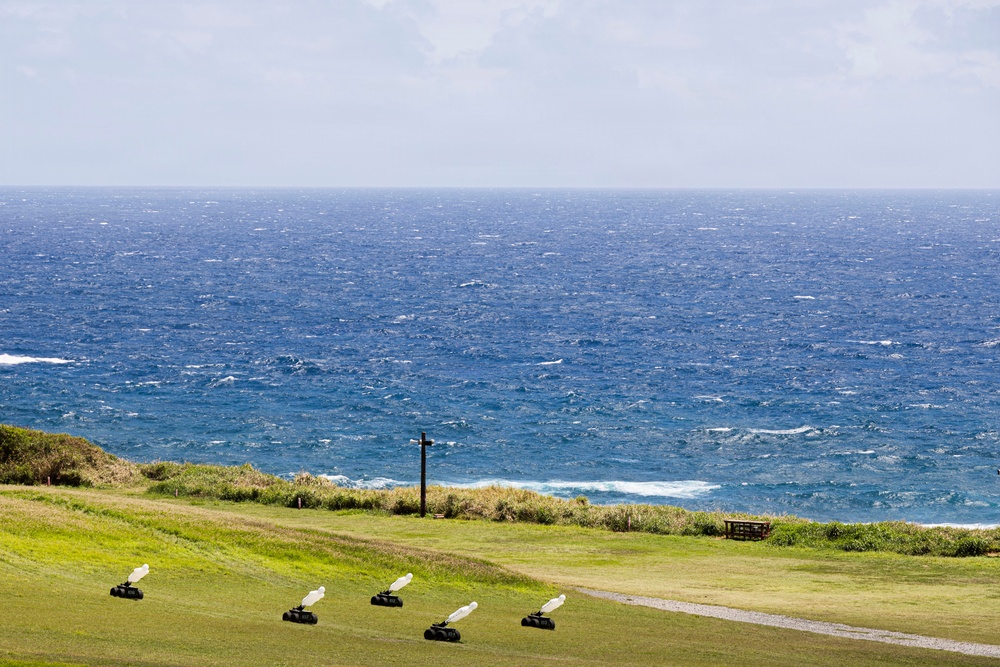 A coastal grassy hillside overlooks a deep blue, choppy ocean under an overcast sky. Four identical small robotic or wheeled devices with white wing-like attachments are arranged in a row on the green lawn. A dark wooden cross or post stands near the cliff edge, and a wooden bench or table is visible to the right.