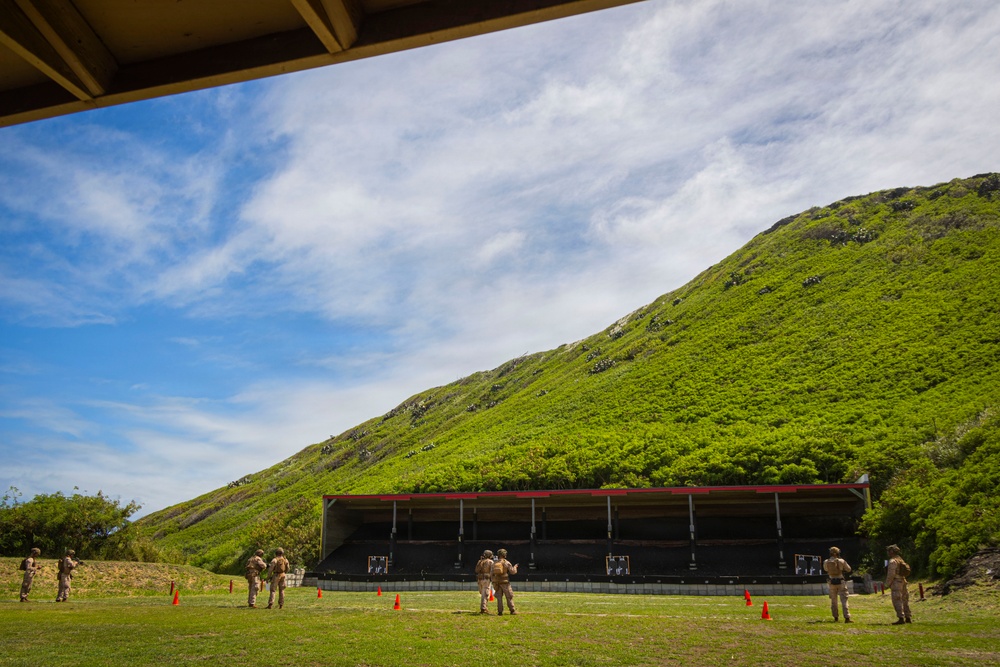 Military personnel in camouflage uniforms are conducting a live-fire weapons qualification exercise at an outdoor shooting range. The range is set against a steep, lush green hillside that serves as a natural backstop. Orange traffic cones mark firing positions along the range line.