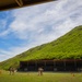 Military personnel in camouflage uniforms are conducting a live-fire weapons qualification exercise at an outdoor shooting range. The range is set against a steep, lush green hillside that serves as a natural backstop. Orange traffic cones mark firing positions along the range line.