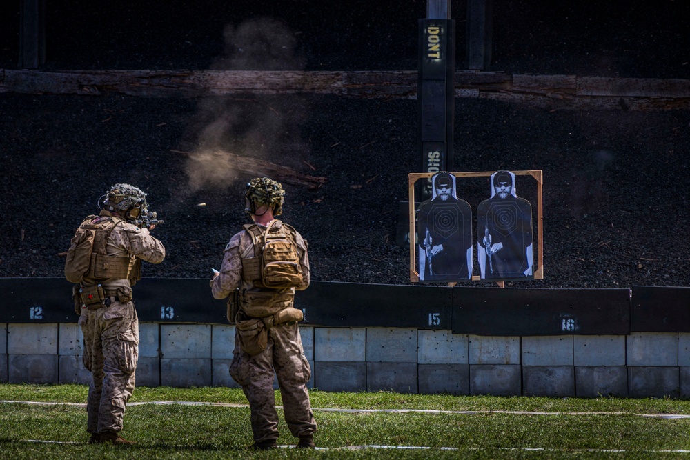 Two military personnel in desert camouflage uniforms are conducting a live-fire shooting exercise at an outdoor range. One soldier is actively firing a rifle at two silhouette paper targets mounted on a wooden frame, while the second appears to be observing or scoring. Visible smoke and a spent casing in mid-air confirm an active discharge.