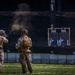 Two military personnel in desert camouflage uniforms are conducting a live-fire shooting exercise at an outdoor range. One soldier is actively firing a rifle at two silhouette paper targets mounted on a wooden frame, while the second appears to be observing or scoring. Visible smoke and a spent casing in mid-air confirm an active discharge.