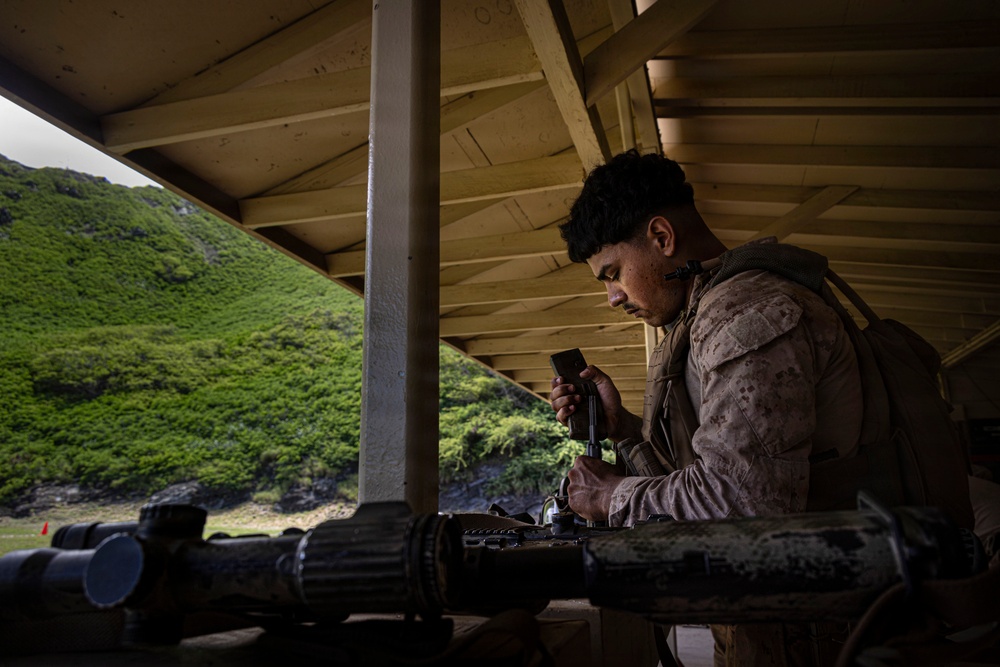 A U.S. Marine in MARPAT camouflage uniform is working on or loading a weapon system under a covered shooting range structure. The setting appears to be a live-fire range with lush green mountainous terrain visible in the background. The scene suggests a training exercise or qualification event at a military range facility.
