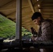 A U.S. Marine in MARPAT camouflage uniform is working on or loading a weapon system under a covered shooting range structure. The setting appears to be a live-fire range with lush green mountainous terrain visible in the background. The scene suggests a training exercise or qualification event at a military range facility.