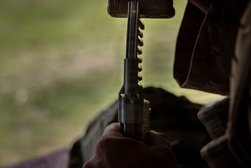 A close-up photograph showing a person holding a firearm, likely a rifle or carbine, with a loaded magazine visible. The image is taken from a low angle with a shallow depth of field, with a blurred green outdoor background. The person appears to be wearing tactical or military-style clothing.
