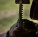 A close-up photograph showing a person holding a firearm, likely a rifle or carbine, with a loaded magazine visible. The image is taken from a low angle with a shallow depth of field, with a blurred green outdoor background. The person appears to be wearing tactical or military-style clothing.