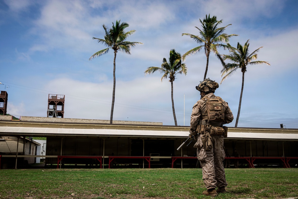 A fully equipped military soldier in desert camouflage (MARPAT pattern) stands on a grass field facing away from the camera, observing a low-profile building with red support columns. The tropical setting is indicated by several tall coconut palm trees and a warm, partly cloudy sky. The scene appears to be a military training exercise or security patrol at a tropical installation.