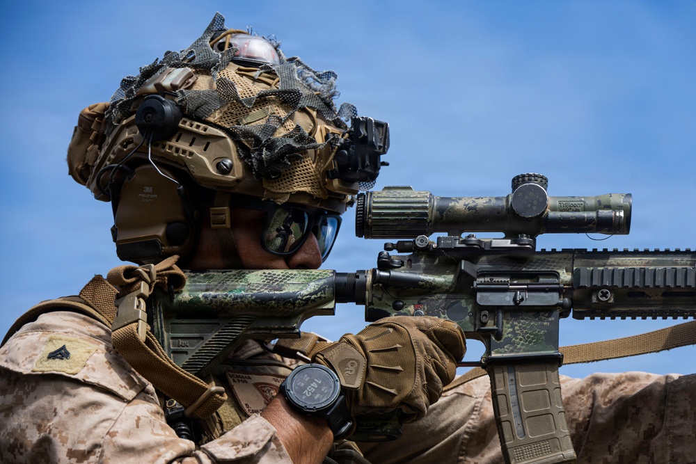 A heavily equipped military sniper is shown in a shooting position, aiming a camouflage-patterned rifle with a large optical scope. The soldier wears desert MARPAT camouflage uniform, a tactical helmet with netting and mounted accessories, and protective eyewear. The image appears to be a professional military photograph taken against a clear blue sky background.
