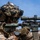 U.S. Marines with 3rd LCT participate in a live-fire basic skills package A heavily equipped military sniper is shown in a shooting position, aiming a camouflage-patterned rifle with a large optical scope. The soldier wears desert MARPAT camouflage uniform, a tactical helmet with netting and mounted accessories, and protective eyewear. The image appears to be a professional military photograph taken against a clear blue sky background.