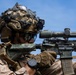 A heavily equipped military sniper is shown in a shooting position, aiming a camouflage-patterned rifle with a large optical scope. The soldier wears desert MARPAT camouflage uniform, a tactical helmet with netting and mounted accessories, and protective eyewear. The image appears to be a professional military photograph taken against a clear blue sky background.