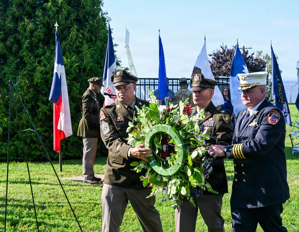 A formal wreath-laying ceremony is taking place outdoors on a sunny day. Three uniformed individuals — two U.S. Army soldiers and one fire department official — are jointly carrying a large floral wreath. Multiple flags, including the Texas state flag, are displayed in the background, suggesting a patriotic or memorial event.