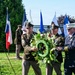 A formal wreath-laying ceremony is taking place outdoors on a sunny day. Three uniformed individuals — two U.S. Army soldiers and one fire department official — are jointly carrying a large floral wreath. Multiple flags, including the Texas state flag, are displayed in the background, suggesting a patriotic or memorial event.