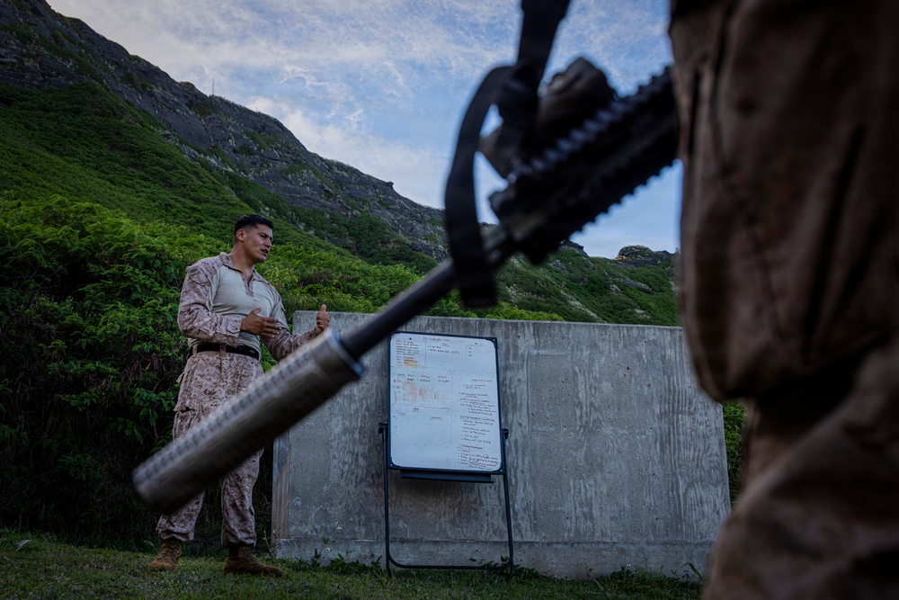 A military service member in Marine Corps MARPAT camouflage uniform is conducting a briefing or instruction session outdoors against a concrete wall, with a whiteboard/easel displaying written notes behind him. The scene is set in a mountainous, heavily vegetated terrain, likely in Hawaii based on the distinctive ridgeline. In the foreground, another service member holding a rifle is visible in shallow focus.