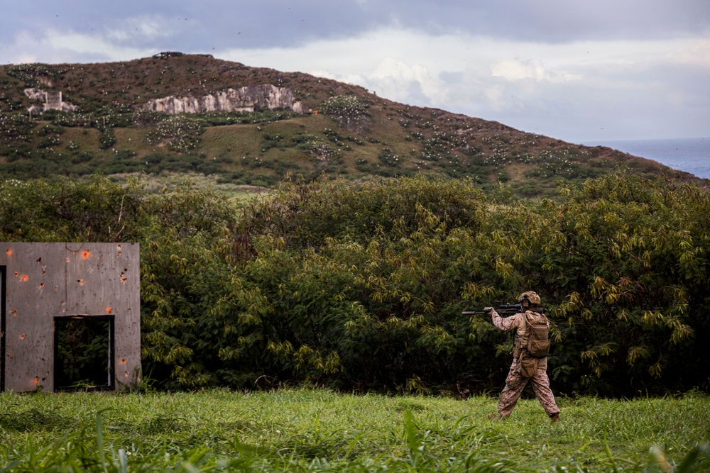 A soldier in desert camouflage uniform is seen in a field aiming or shouldering a weapon system, positioned near a bullet-riddled metal structure used as a training target. The setting appears to be a military training range on a tropical or subtropical island, with dense vegetation, rocky hillside, and ocean visible in the background. The scene is consistent with a live-fire training exercise.