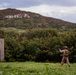 A soldier in desert camouflage uniform is seen in a field aiming or shouldering a weapon system, positioned near a bullet-riddled metal structure used as a training target. The setting appears to be a military training range on a tropical or subtropical island, with dense vegetation, rocky hillside, and ocean visible in the background. The scene is consistent with a live-fire training exercise.