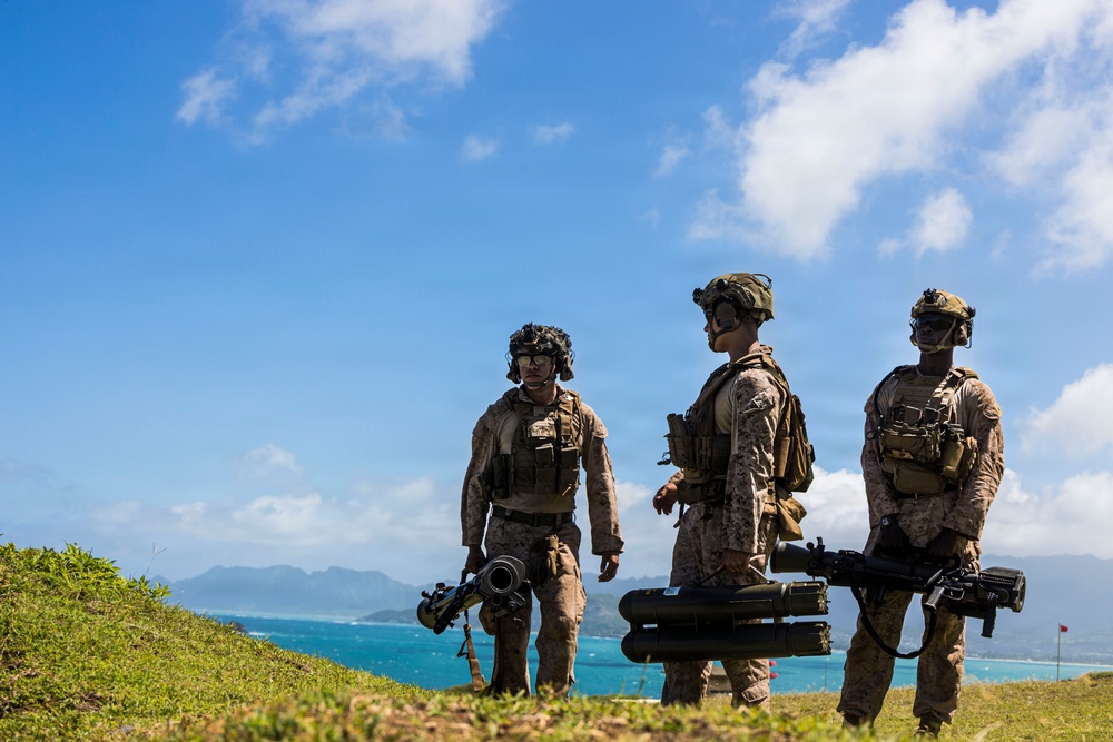 Three military personnel in full combat gear are positioned on a grassy elevated terrain overlooking a turquoise coastal bay with mountains in the background. They are equipped with tactical vests, helmets with night-vision mounts, and are carrying what appear to be shoulder-fired rocket/missile launchers. The setting appears to be a tropical island location based on the vegetation, water color, and mountain terrain.