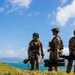 Three military personnel in full combat gear are positioned on a grassy elevated terrain overlooking a turquoise coastal bay with mountains in the background. They are equipped with tactical vests, helmets with night-vision mounts, and are carrying what appear to be shoulder-fired rocket/missile launchers. The setting appears to be a tropical island location based on the vegetation, water color, and mountain terrain.