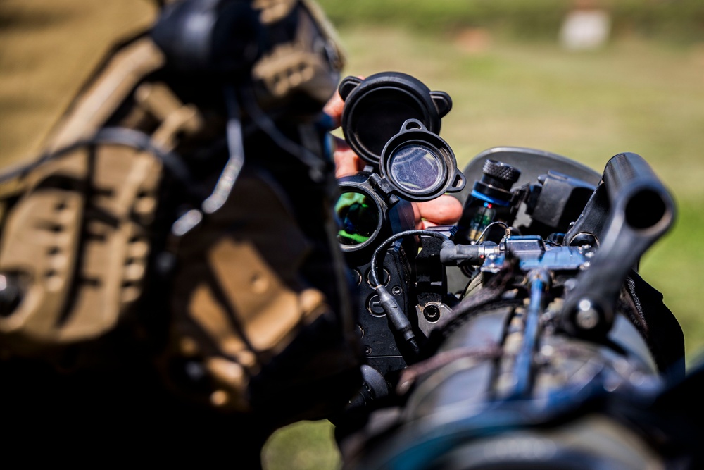 A close-up photograph of a military or tactical operator aiming through an optical scope mounted on a weapon system. The image is shot with shallow depth of field, keeping the optic and operator's hands in focus while the background and operator's body remain blurred. The equipment appears to be heavily accessorized with multiple attachments.
