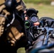 A close-up photograph of a military or tactical operator aiming through an optical scope mounted on a weapon system. The image is shot with shallow depth of field, keeping the optic and operator's hands in focus while the background and operator's body remain blurred. The equipment appears to be heavily accessorized with multiple attachments.