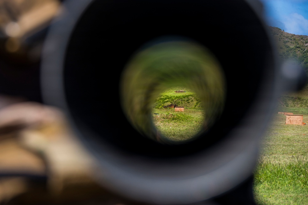 The image is taken through the eyepiece of a rifle scope at what appears to be an outdoor shooting range. The scope's circular field of view reveals a grassy downrange area with earthen berms and wooden target boxes. The surrounding environment outside the scope shows a green hillside under a partly cloudy sky.