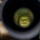 U.S. Marines with 3rd LCT participate in a live-fire basic skills package The image is taken through the eyepiece of a rifle scope at what appears to be an outdoor shooting range. The scope's circular field of view reveals a grassy downrange area with earthen berms and wooden target boxes. The surrounding environment outside the scope shows a green hillside under a partly cloudy sky.