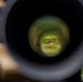 The image is taken through the eyepiece of a rifle scope at what appears to be an outdoor shooting range. The scope's circular field of view reveals a grassy downrange area with earthen berms and wooden target boxes. The surrounding environment outside the scope shows a green hillside under a partly cloudy sky.