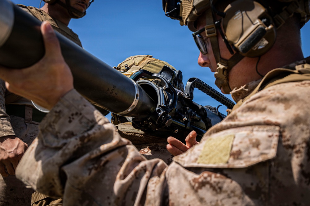 Two military personnel in desert MARPAT camouflage uniforms are handling what appears to be a crew-served weapon system, likely an M3 Carl Gustaf recoilless rifle or similar anti-armor weapon. The soldier on the right is wearing a combat helmet with communications headset and protective goggles. The image captures a close-up training or operational scenario under clear blue sky conditions.