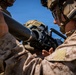 Two military personnel in desert MARPAT camouflage uniforms are handling what appears to be a crew-served weapon system, likely an M3 Carl Gustaf recoilless rifle or similar anti-armor weapon. The soldier on the right is wearing a combat helmet with communications headset and protective goggles. The image captures a close-up training or operational scenario under clear blue sky conditions.