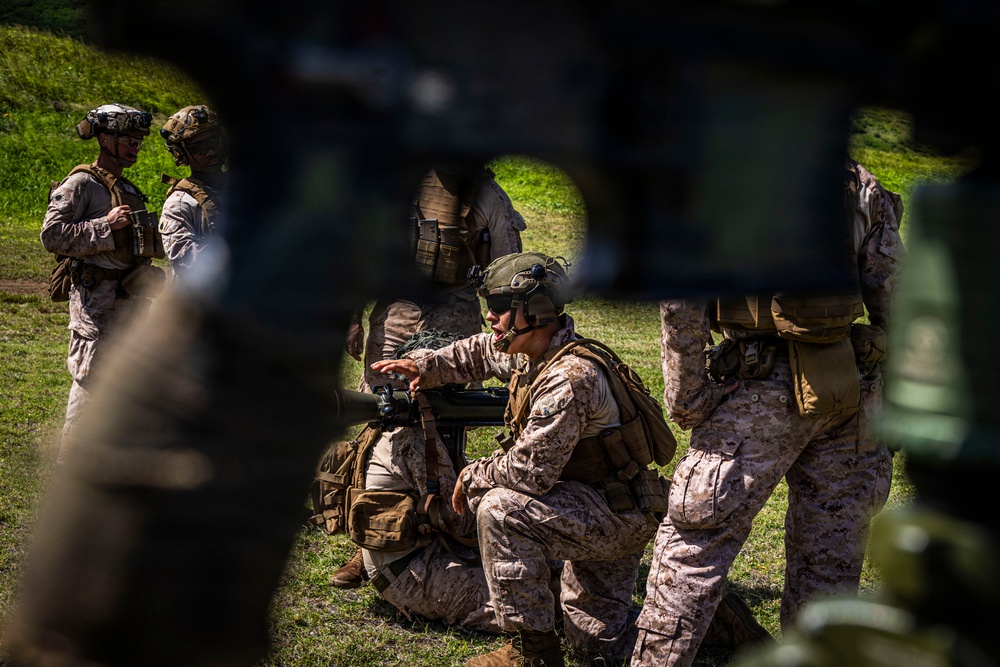 The image shows a group of military personnel in desert camouflage uniforms (MARPAT pattern) conducting what appears to be a field training exercise or operational briefing in an outdoor grassy area. A central figure is crouched near equipment, appearing to operate or demonstrate a piece of gear while others observe. The photo is taken through or around foreground obstructions, creating a partially obscured perspective.