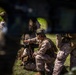The image shows a group of military personnel in desert camouflage uniforms (MARPAT pattern) conducting what appears to be a field training exercise or operational briefing in an outdoor grassy area. A central figure is crouched near equipment, appearing to operate or demonstrate a piece of gear while others observe. The photo is taken through or around foreground obstructions, creating a partially obscured perspective.