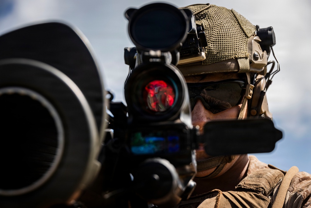 A military operator is shown in close-up, aiming a weapon system equipped with an optical sight displaying a red reticle. The individual is wearing full tactical gear including a ballistic helmet with mounted accessories, protective eyewear, and hearing protection. The image is a high-quality professional photograph likely taken during a training exercise or operational deployment.