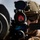 U.S. Marines with 3rd LCT participate in a live-fire basic skills package A military operator is shown in close-up, aiming a weapon system equipped with an optical sight displaying a red reticle. The individual is wearing full tactical gear including a ballistic helmet with mounted accessories, protective eyewear, and hearing protection. The image is a high-quality professional photograph likely taken during a training exercise or operational deployment.