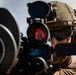 A military operator is shown in close-up, aiming a weapon system equipped with an optical sight displaying a red reticle. The individual is wearing full tactical gear including a ballistic helmet with mounted accessories, protective eyewear, and hearing protection. The image is a high-quality professional photograph likely taken during a training exercise or operational deployment.