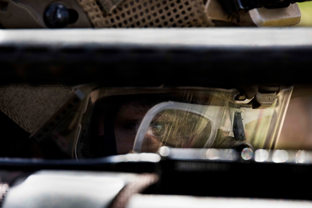 A person wearing military or tactical gear, including a helmet and protective eyewear/goggles, is visible through a narrow gap between vehicle panels or armor plating. The image is taken from outside a military-style vehicle, looking through what appears to be a window or hatch opening. The composition creates a framed, confined view of the individual inside.