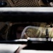 A person wearing military or tactical gear, including a helmet and protective eyewear/goggles, is visible through a narrow gap between vehicle panels or armor plating. The image is taken from outside a military-style vehicle, looking through what appears to be a window or hatch opening. The composition creates a framed, confined view of the individual inside.