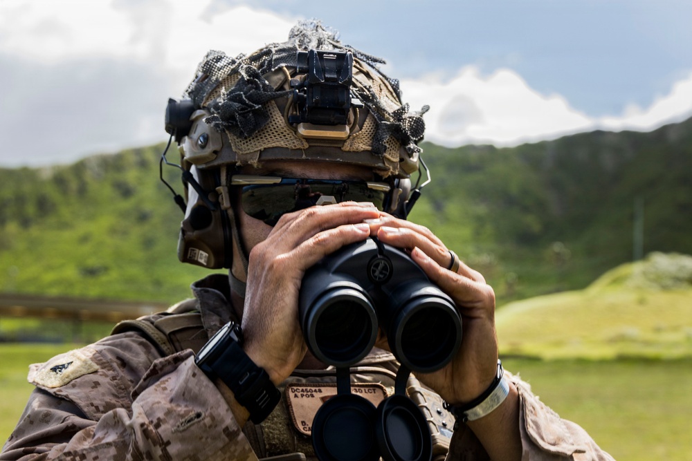 A military service member in desert MARPAT camouflage uniform is using binoculars to observe a distant area. The individual is wearing a tactical helmet with a camouflage cover, night vision device mount, and communications equipment. The setting appears to be a lush, green hilly or mountainous outdoor environment under partly cloudy skies.