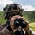 U.S. Marines with 3rd LCT participate in a live-fire basic skills package A military service member in desert MARPAT camouflage uniform is using binoculars to observe a distant area. The individual is wearing a tactical helmet with a camouflage cover, night vision device mount, and communications equipment. The setting appears to be a lush, green hilly or mountainous outdoor environment under partly cloudy skies.